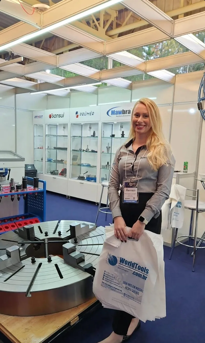 Atendente da World Tools com sacolas da marca ao lado da placa de torno gigante na Intermach 2025